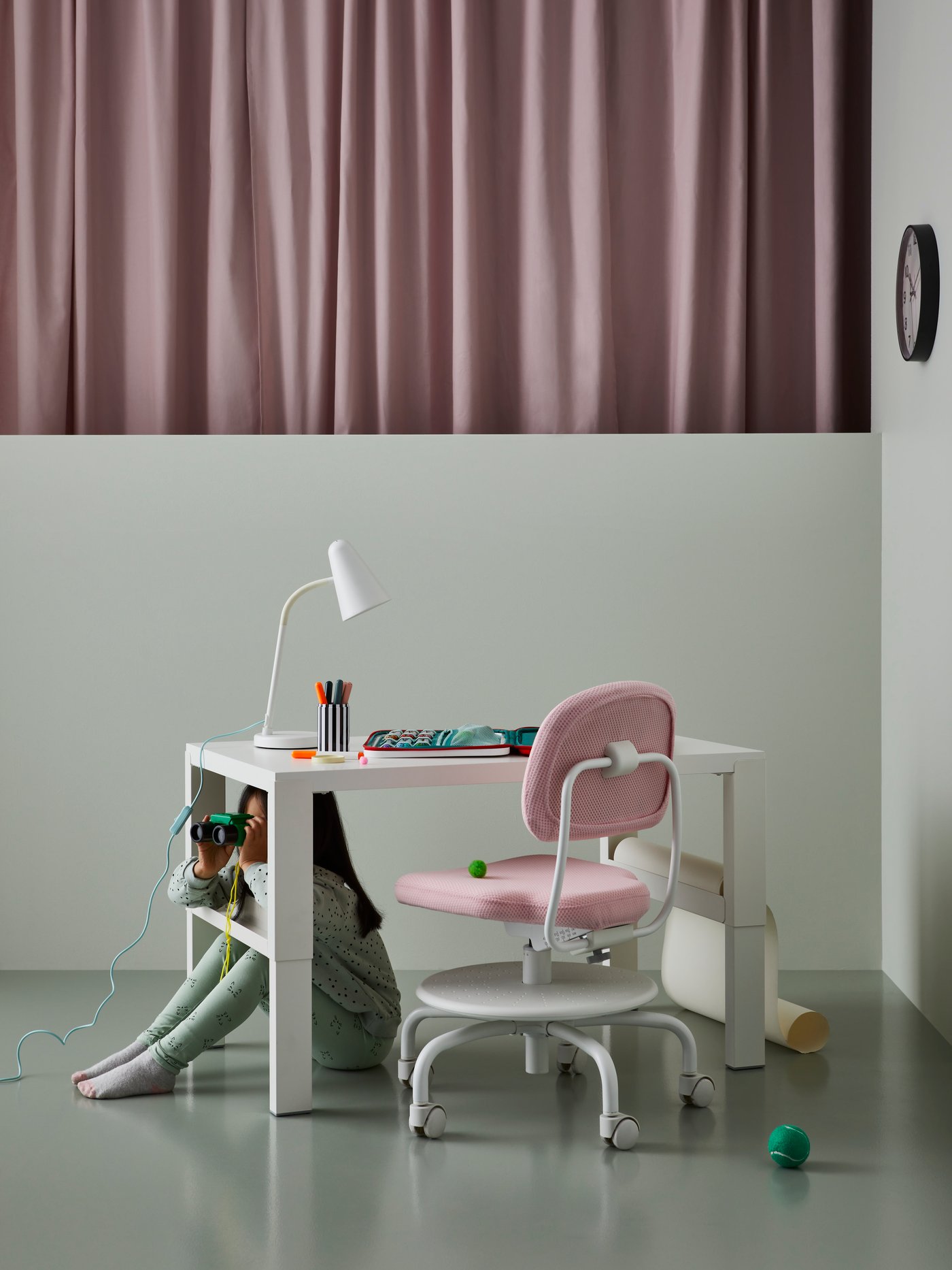 A child sits under a PÅHL desk with a work lamp and art supplies on it. A pink children’s desk chair is pushed underneath it.