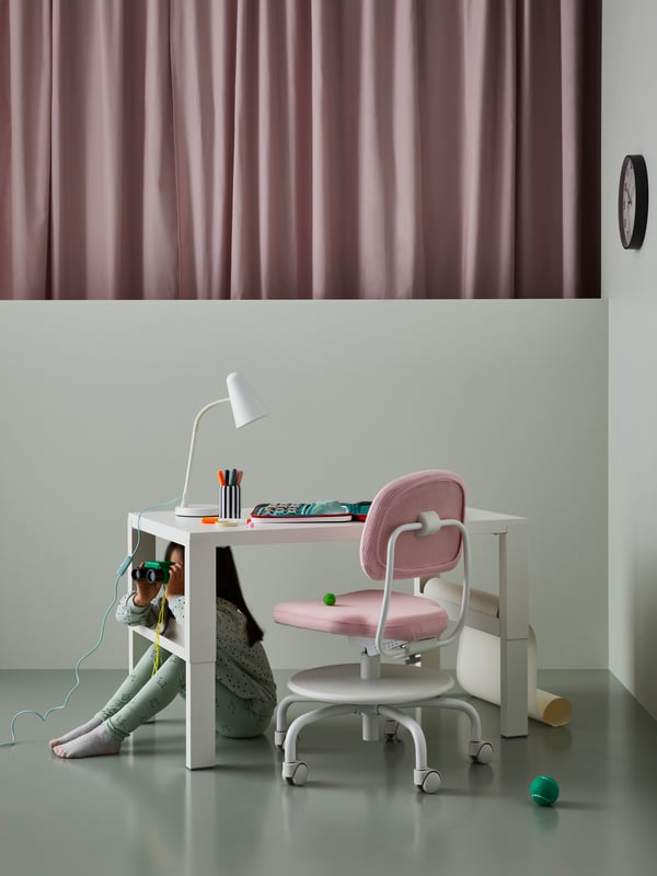 A child sits under a PÅHL desk with a work lamp and art supplies on it. A pink children’s desk chair is pushed underneath it.