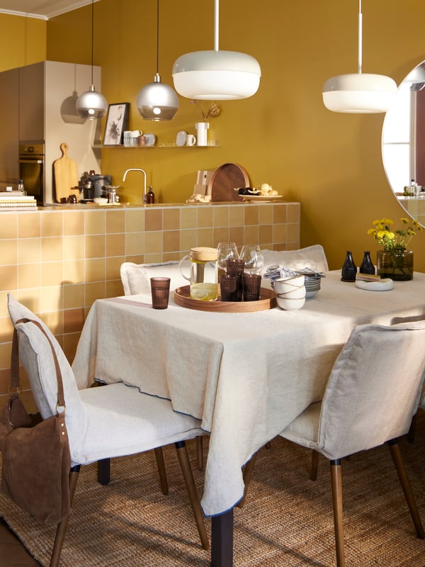 A room with a dining table with a tablecloth, KLINTEN chairs in light beige and BLÅSVERK pendant lamps in beige hanging.