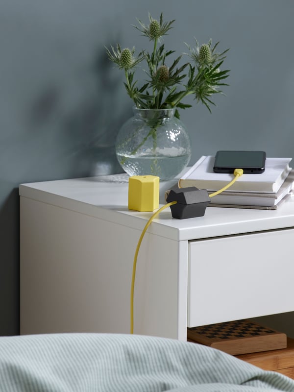 A black HAVSKÅL USB anchor sits on a bedside table, holding a yellow phone charging cable attached to a black mobile phone.