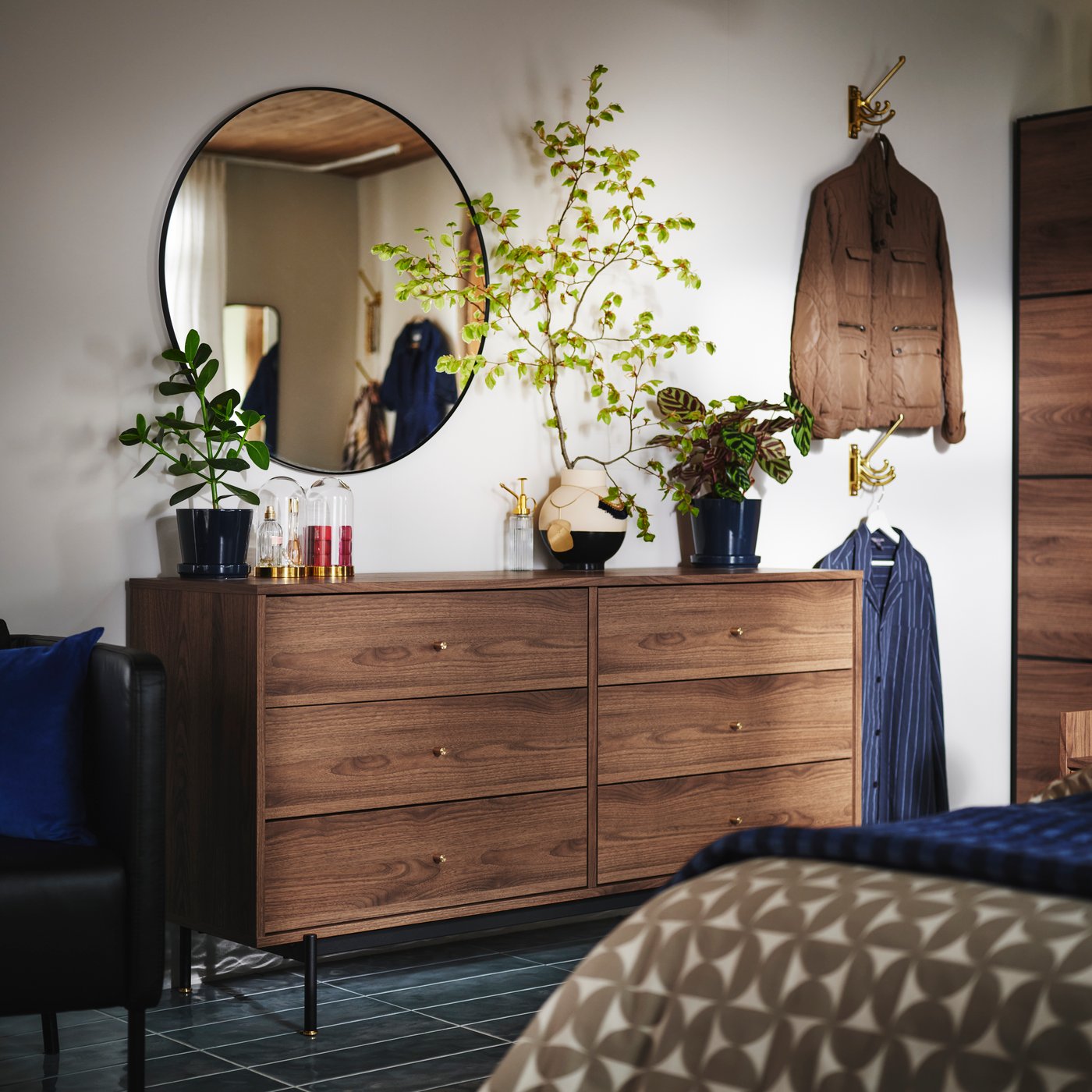 A brown walnut-effect RÅDMANSÖ chest of 6 drawers has a round LYNDBYN mirror above it and plants and other items on top.