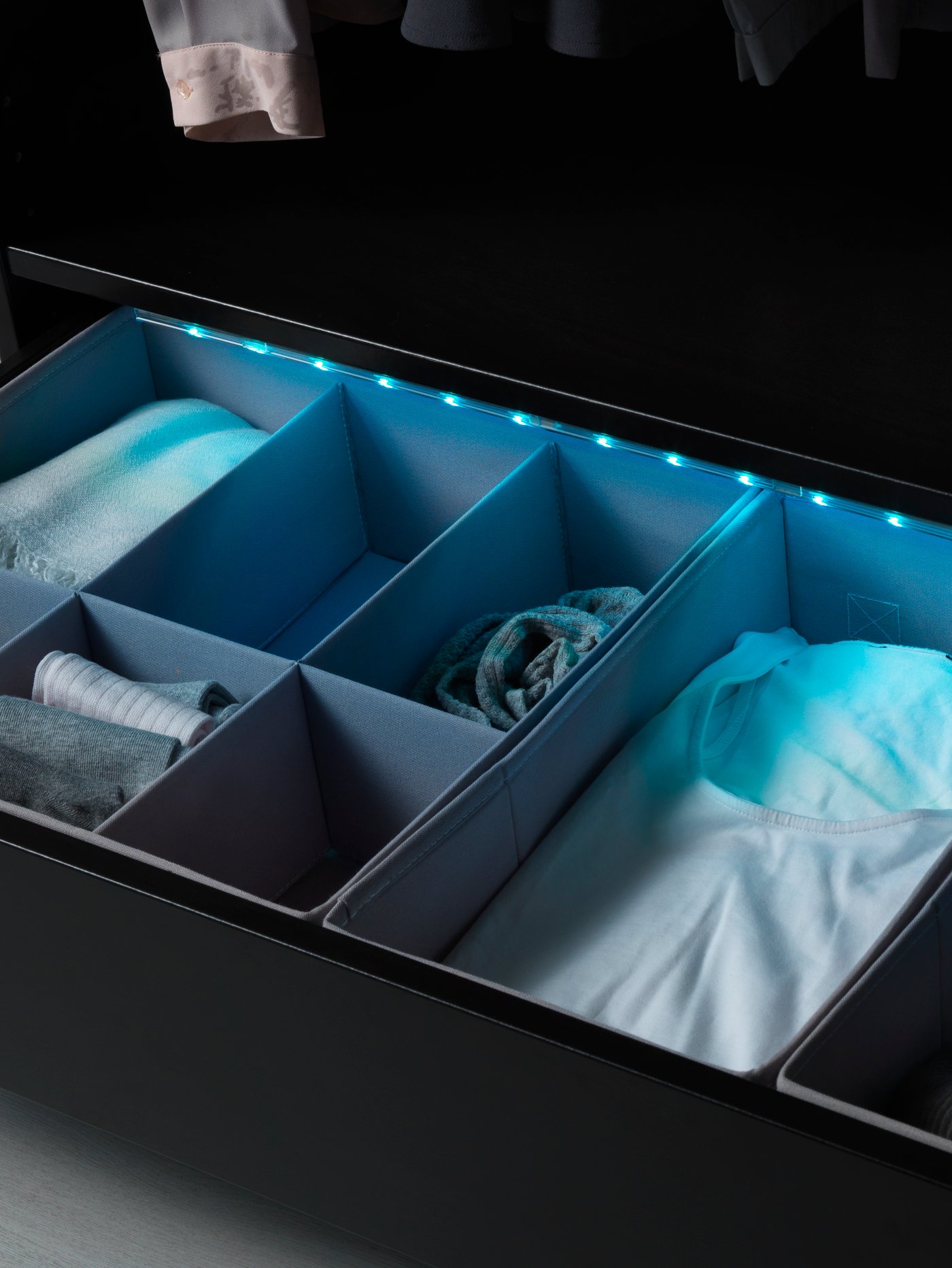 A wardrobe drawer fitted with organisers and filled clothes and accessories. The drawer is lit by a blue LED light strip.