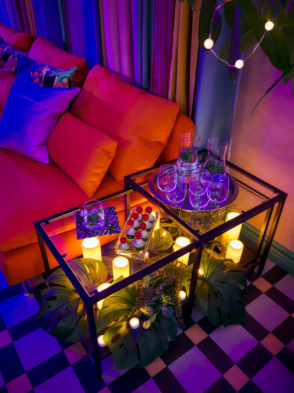 A VITTSJÖ nest of tables. Snacks on top, a carpet of FEJKA and SMYCKA artificial leaves and GODAFTON LED block candles below.