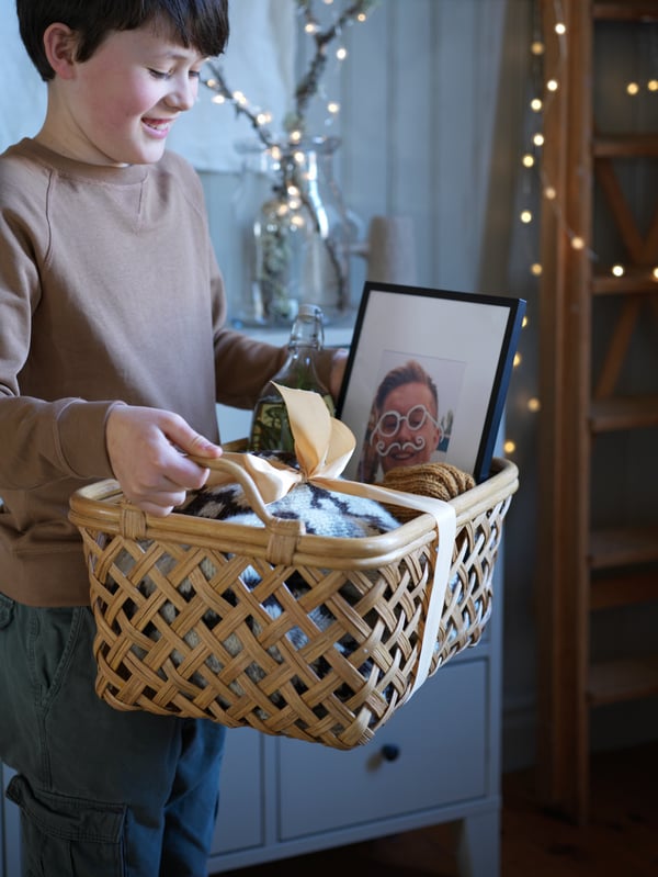 Ein Kind halt einen INSVEP Rattankorb mit Griffen, der mit festlichen Geschenken gefüllt ist, u. a. einem LOMVIKEN schwarzen Rahmen.