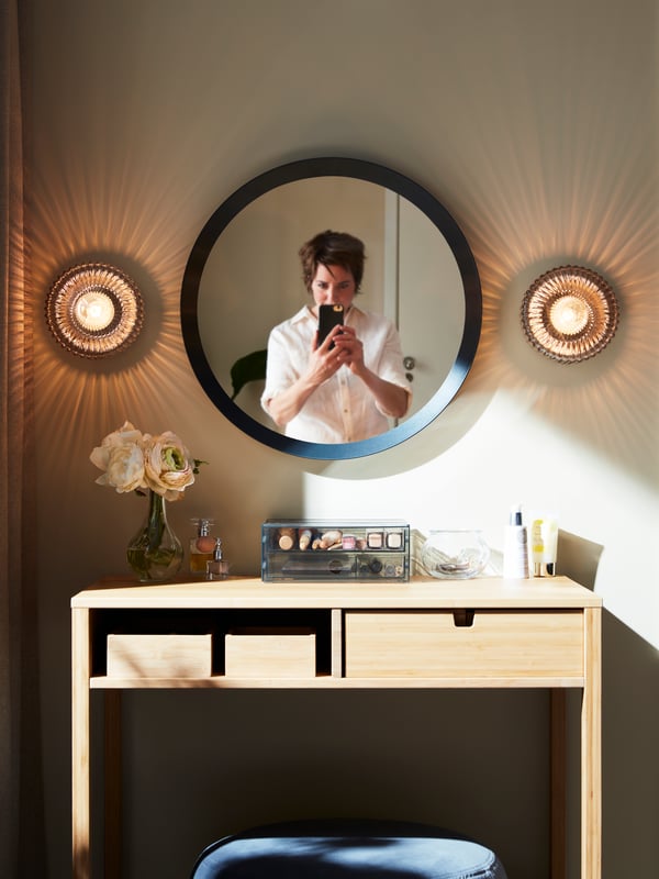 Woman in a white shirt taking a picture in front of a mirror with brass/grey clear glass SOLKLINT wall lamps on either side.