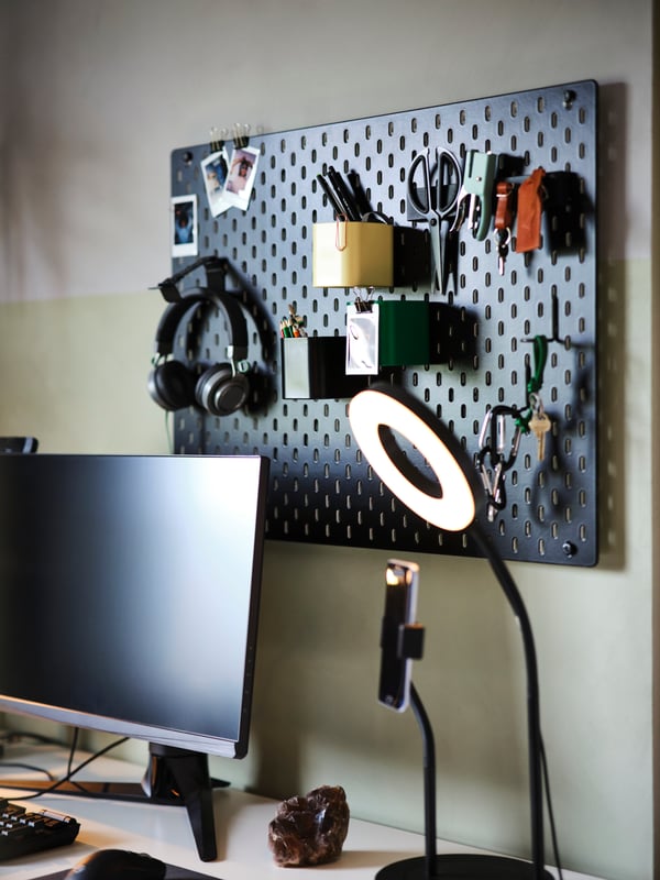 A black SKÅDIS pegboard with headphones and accessories hang over a desk with a screen and a LÅNESPELARE ring light on it.