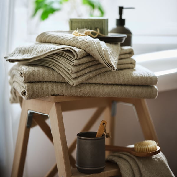 A pile of VÅGSJÖN cotton towels in beige colour on a wooden stool in front of a window. Soap and a body brush next to it.
