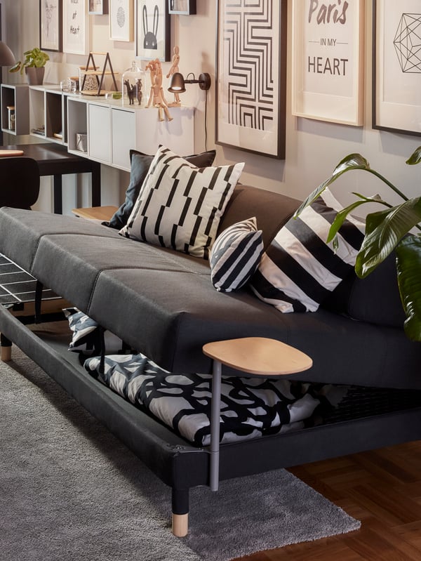 A dark grey FLOTTEBO sofa-bed where the seat has been lifted, underneath it is storage space with a duvet and a pillow.