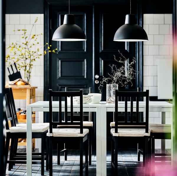 Two black SKURUP pendant lamps hang over two white MELLTORP tables which have some brown-black STEFAN chairs around them.