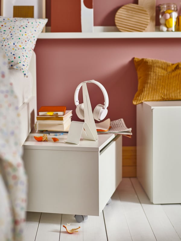 A white SLÄKT storage box with castors beside a bed, working as a bedside table, with a headset/tablet stand on top.