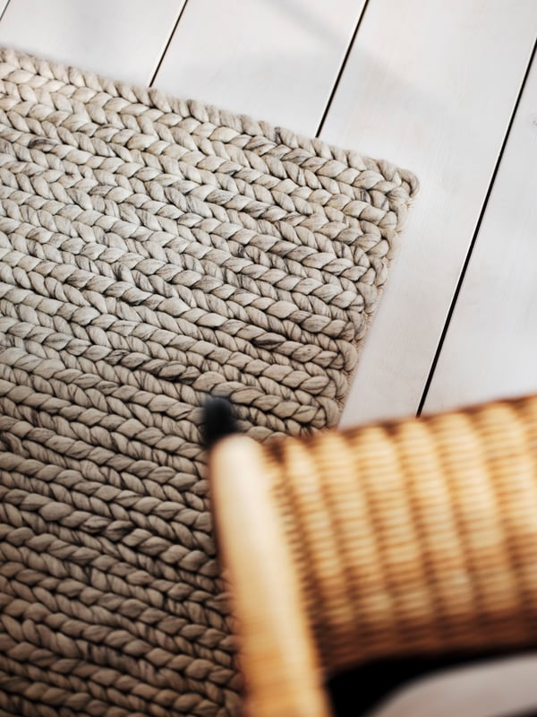 A white, wooden floor with a handmade/off-white HJORTSVANG rug and the corner of a rattan/anthracite ULRIKSBERG armchair.