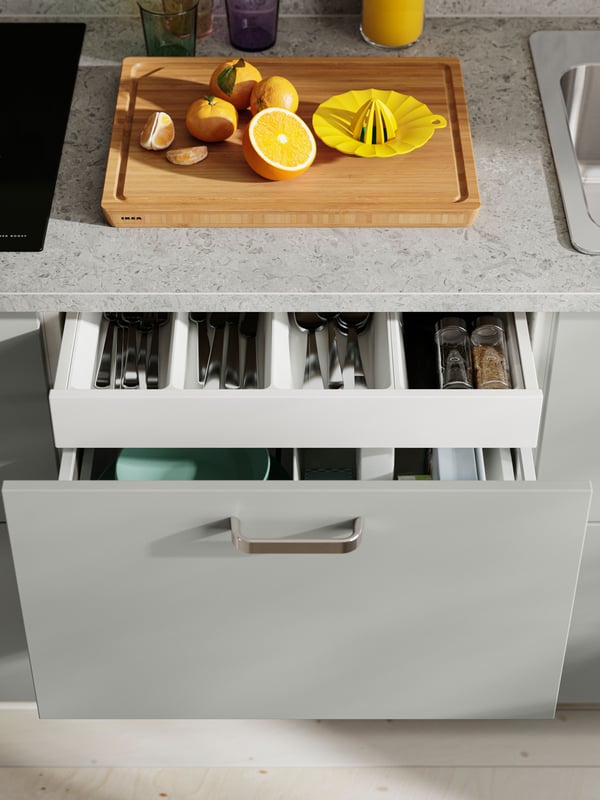 A drawer with HAVSTORP light grey cabinet doors with organised utensils, with a cutting board and a citrus juicer on the countertop.