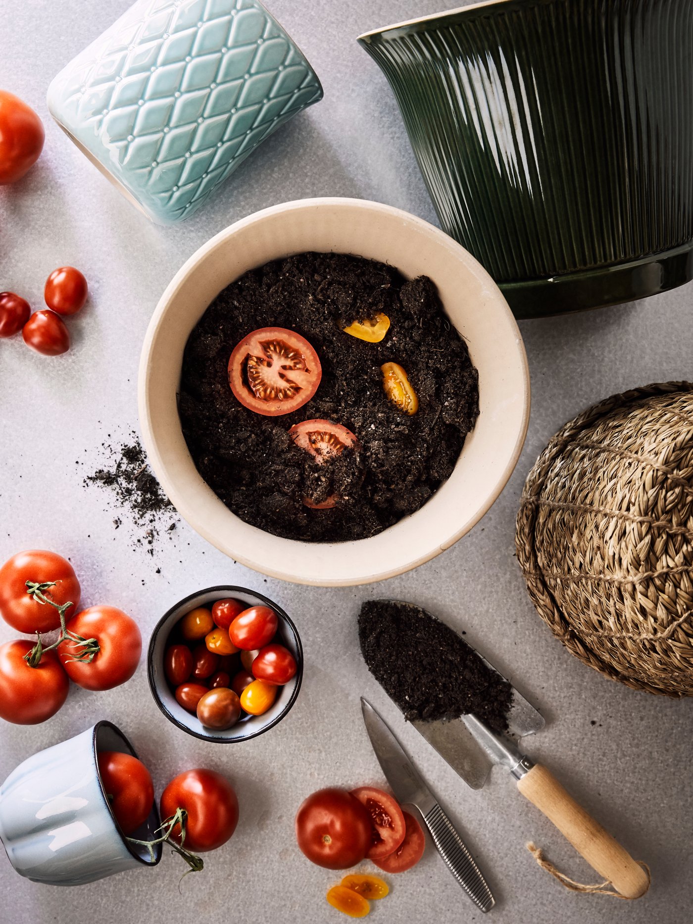Verschiedene Übertöpfe, u. a. KAMOMILL Übertopf in Grün, reife Tomaten und Scheiben gelber und roter Tomaten, die in frische Erde gelegt wurden.