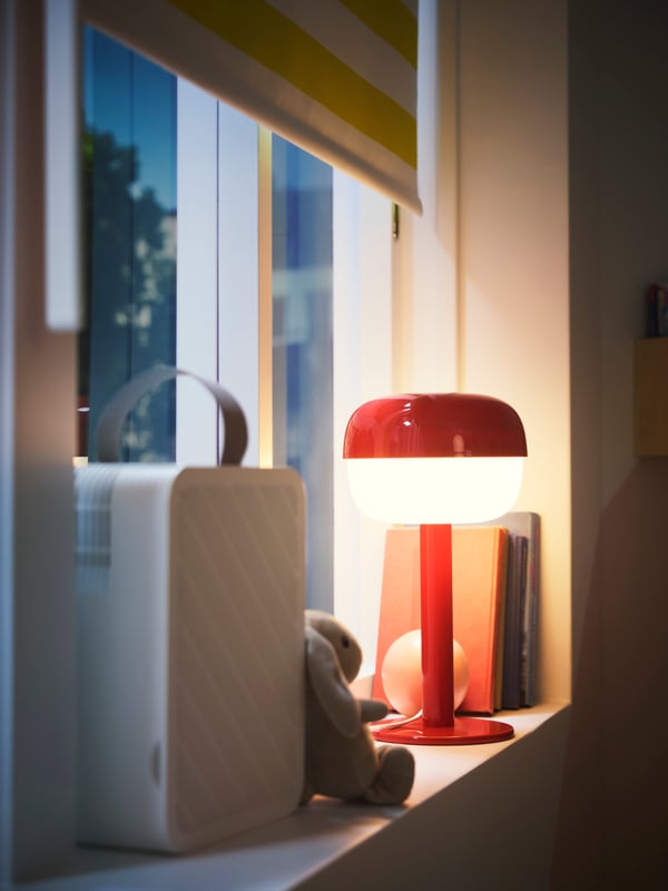 Närbild av ett fönster med en mörkläggande rullgardin, en UPPÅTVIND luftrenare och en bordslampa som står på fönsterbrädan.