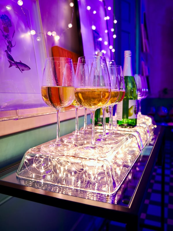 Inverted MIXTUR glass oven dishes with VISSVASS lighting chains underneath, forming a glowing stand for filled wine glasses.
