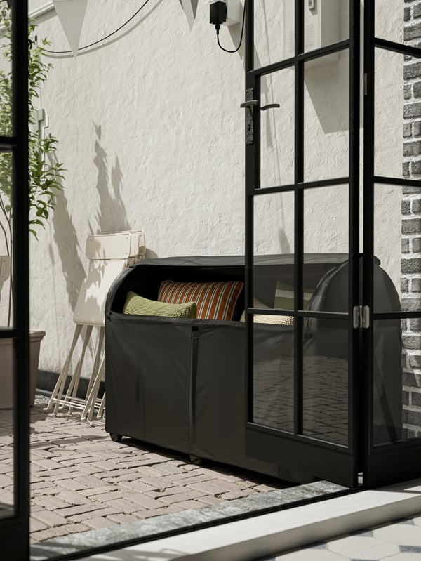 A large black outdoor storage box with green and orange cushions against a white wall, beside an open glass door.