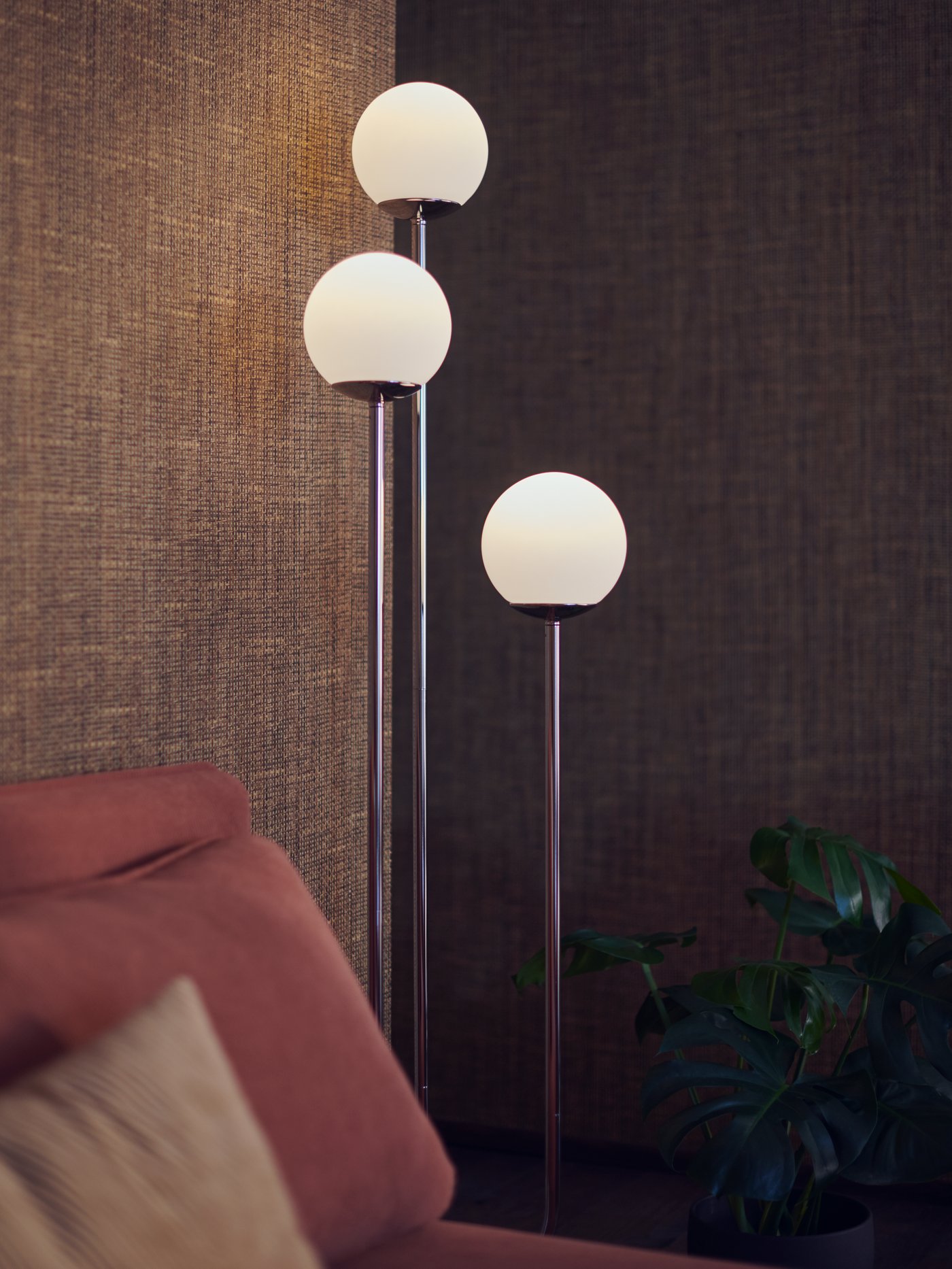 A SIMRISHAMN floor lamp in the corner of a living room with hessian wall covering, a rust sofa and a leafy green plant.