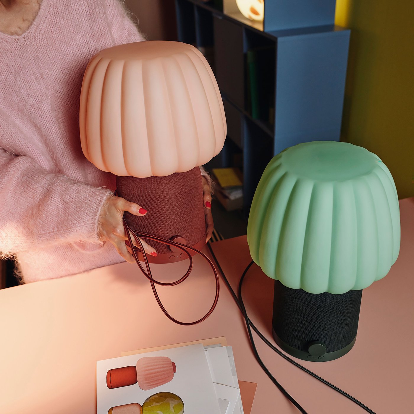 Två KULGLASS bordslampor/högtalare med veckade skärmar, en rosa och en grön, står på ett rosa bord. En person håller i den rosa lampan, och en broschyr med lampbilder ligger bredvid.