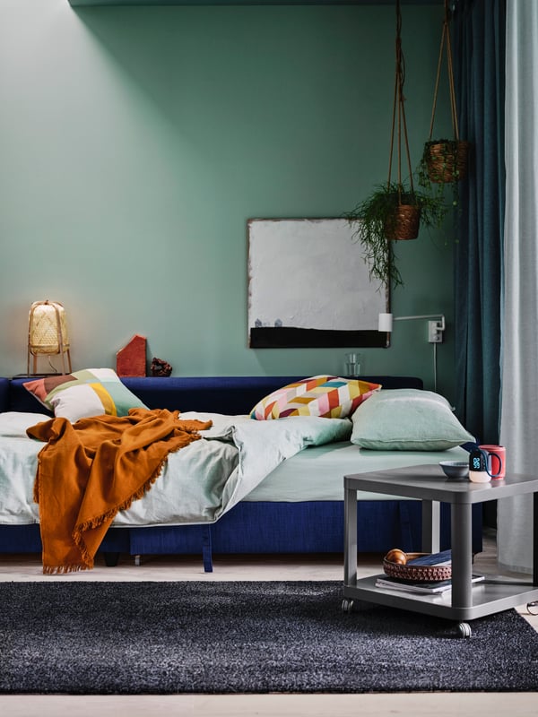A room with light-green walls, a blue FRIHETEN corner sofa-bed with bed linen and textiles, and a grey TINGBY side table.