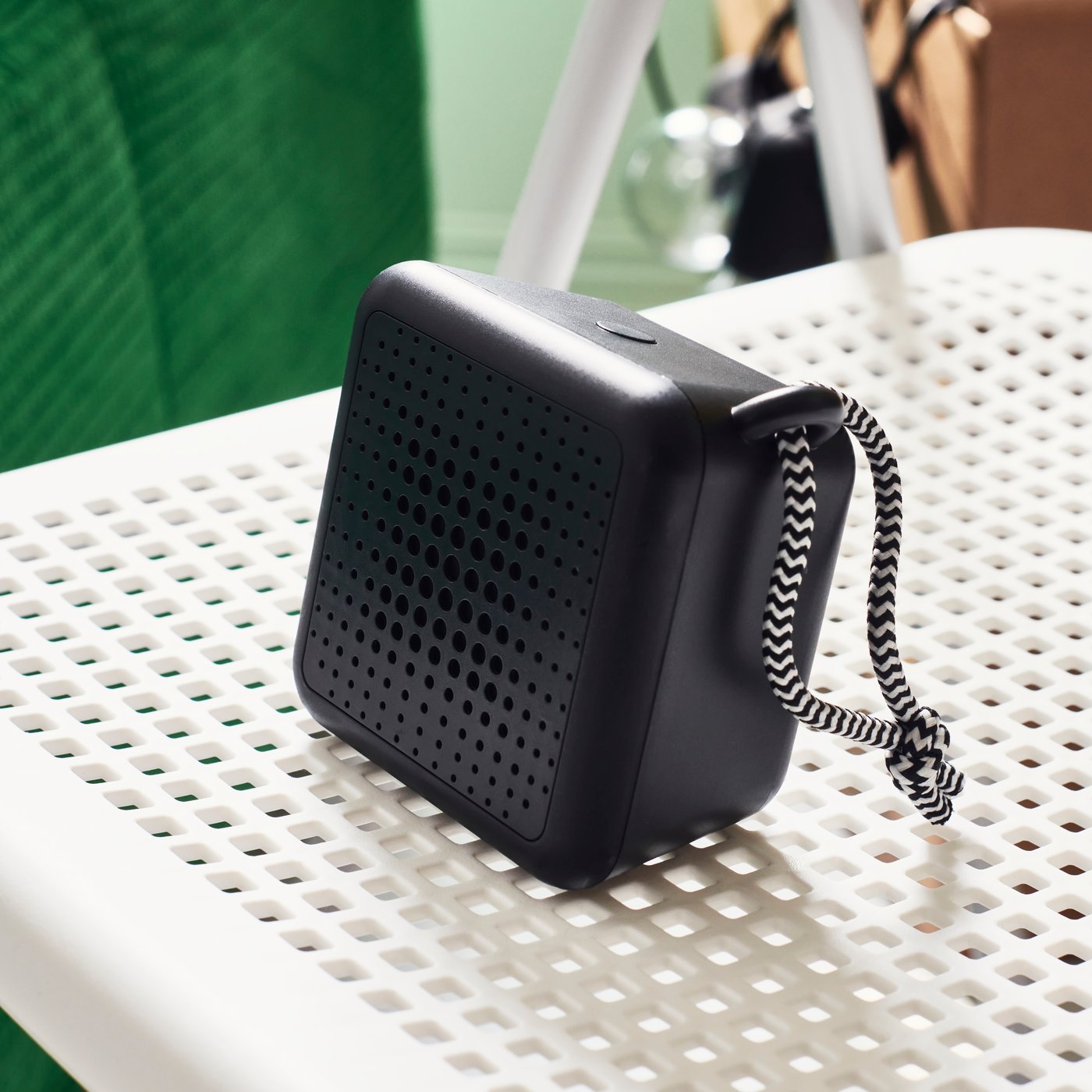 A black portable speaker sits atop a white, perforated flat surface.  It has a black and white striped cord handle.