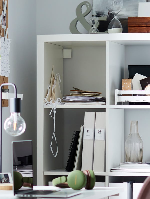 A white KALLAX shelving unit attached to the wall next to a desk with a laptop, and various items on the shelves.