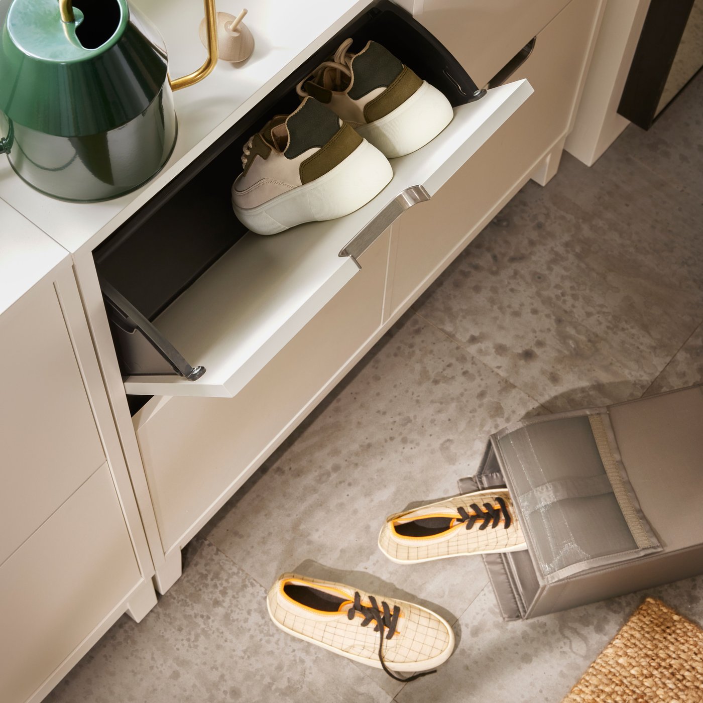In a room is a white closed shoe storage. One drawer is open with sneakers in it. On the drawer is a green watering can.