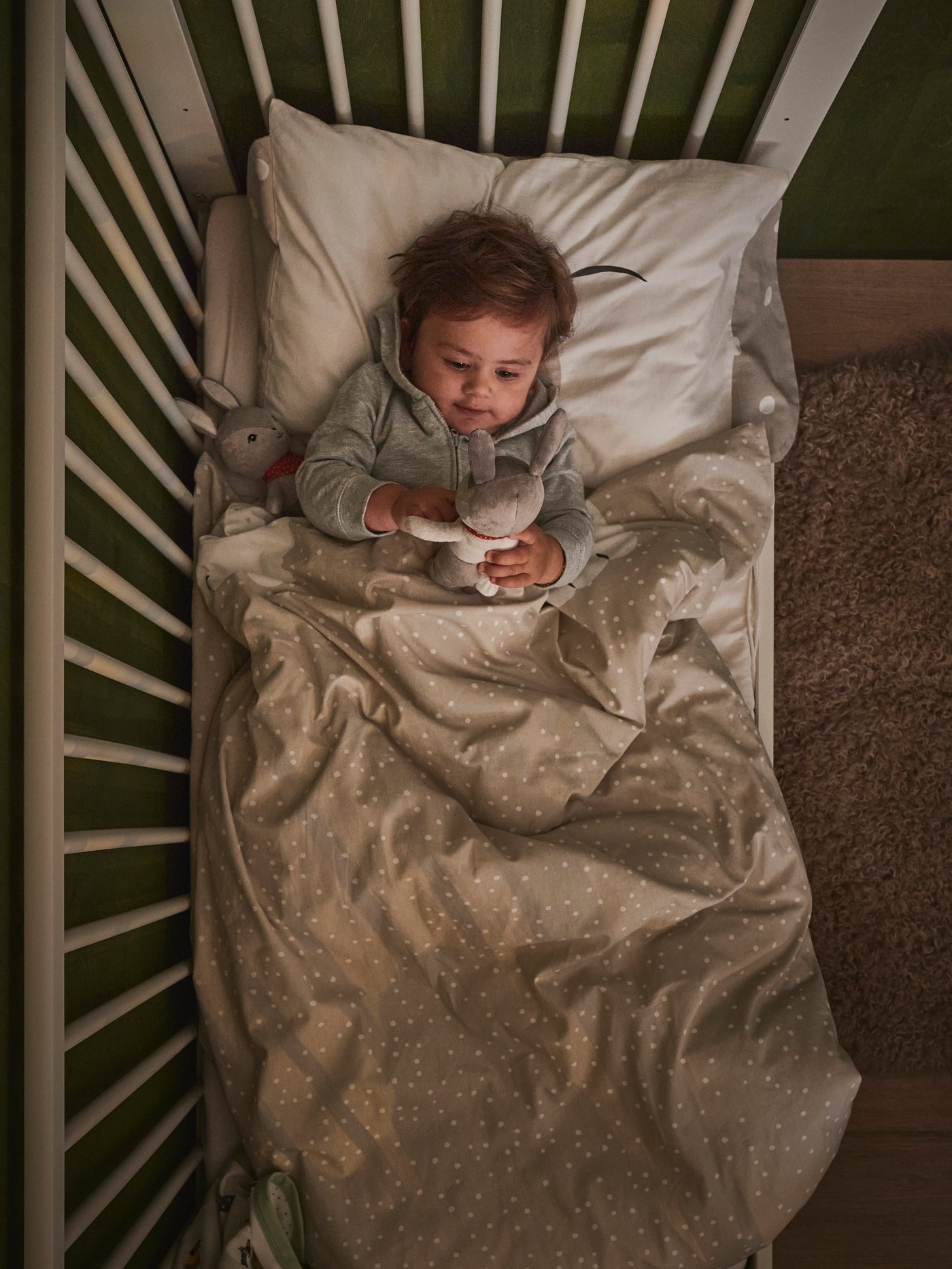 A child holding a soft toy lying on their back in a white GULLIVER cot with one side removed, covered in DRÖMSLOTT bed linen.