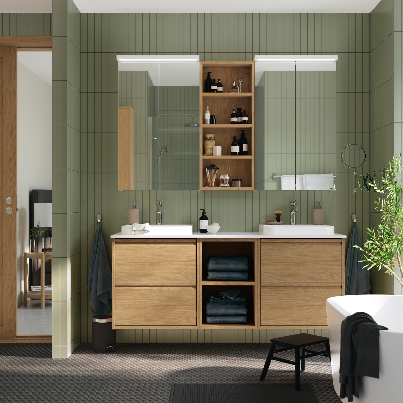 A bathroom with green tiles and wooden ÄNGSJÖN bathroom furniture, featuring mirrored cabinets and an open shelving unit above.