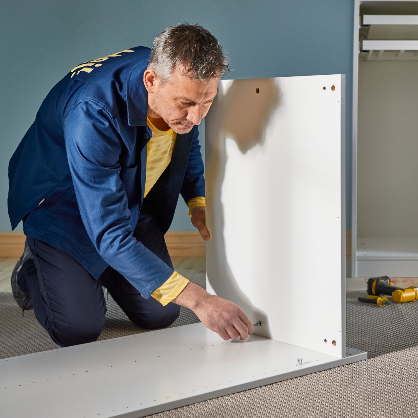 A coworker, clad in a blue work overall, is holding and tightening the inside bracket of a white table leg.