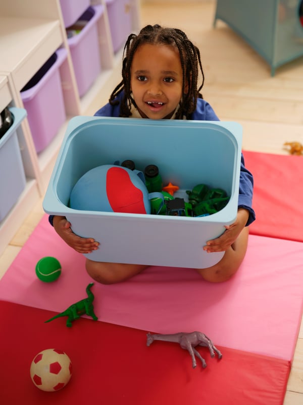 A child sitting on a pink and red PLUFSIG foldable gym mat, holding a light blue TROFAST storage box with toys in it.