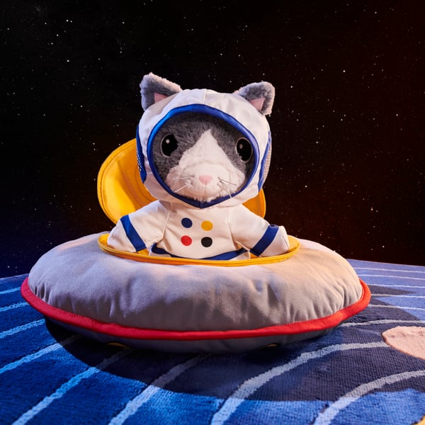 An AFTONSPARV soft toy cat wearing an astronaut suit and sitting in an AFTONSPARV soft toy spaceship on top of a space rug.
