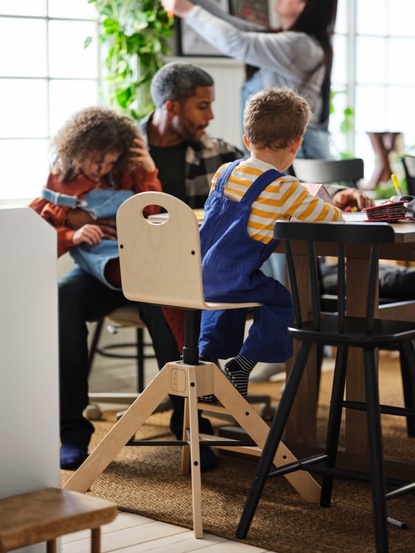 Four family members in a dining area. One child sits on a GRÅVAL birch highchair standing next to a black AGAM junior chair.