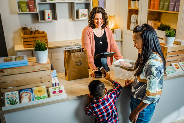 A store checkout where a person pays with a card at a card terminal, while another person stands behind the counter and a child leans against the edge.