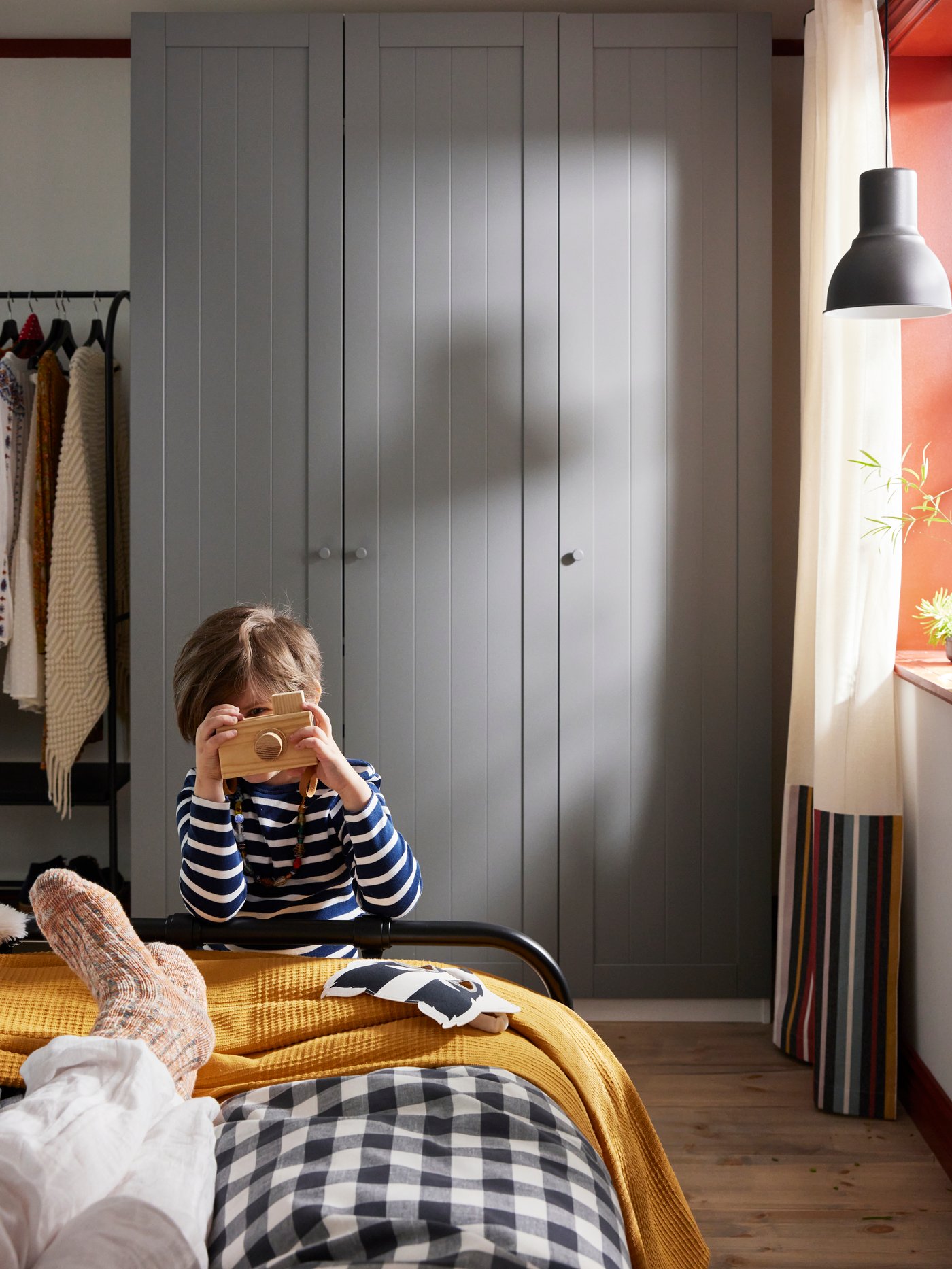 The foot-end of a bed and grey PAX wardrobe behind with open storage next to them. A child takes photo with toy camera.