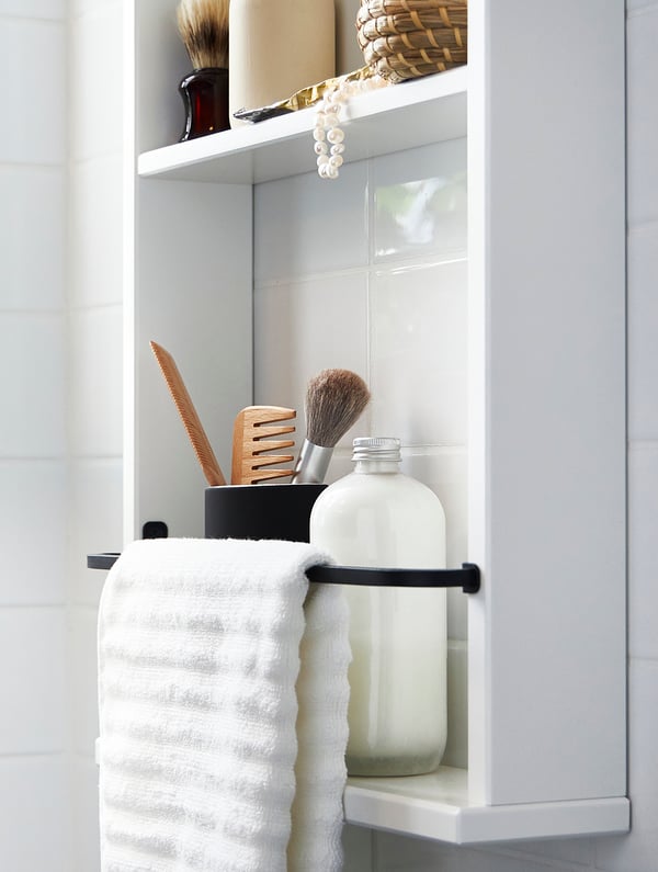 Un estante HEMNES blanco con una toalla blanca colgada sobre una baranda negra. Sobre la repisa se ve una botella y un recipiente con productos de baño como peinetas y un pincel de maquillaje.