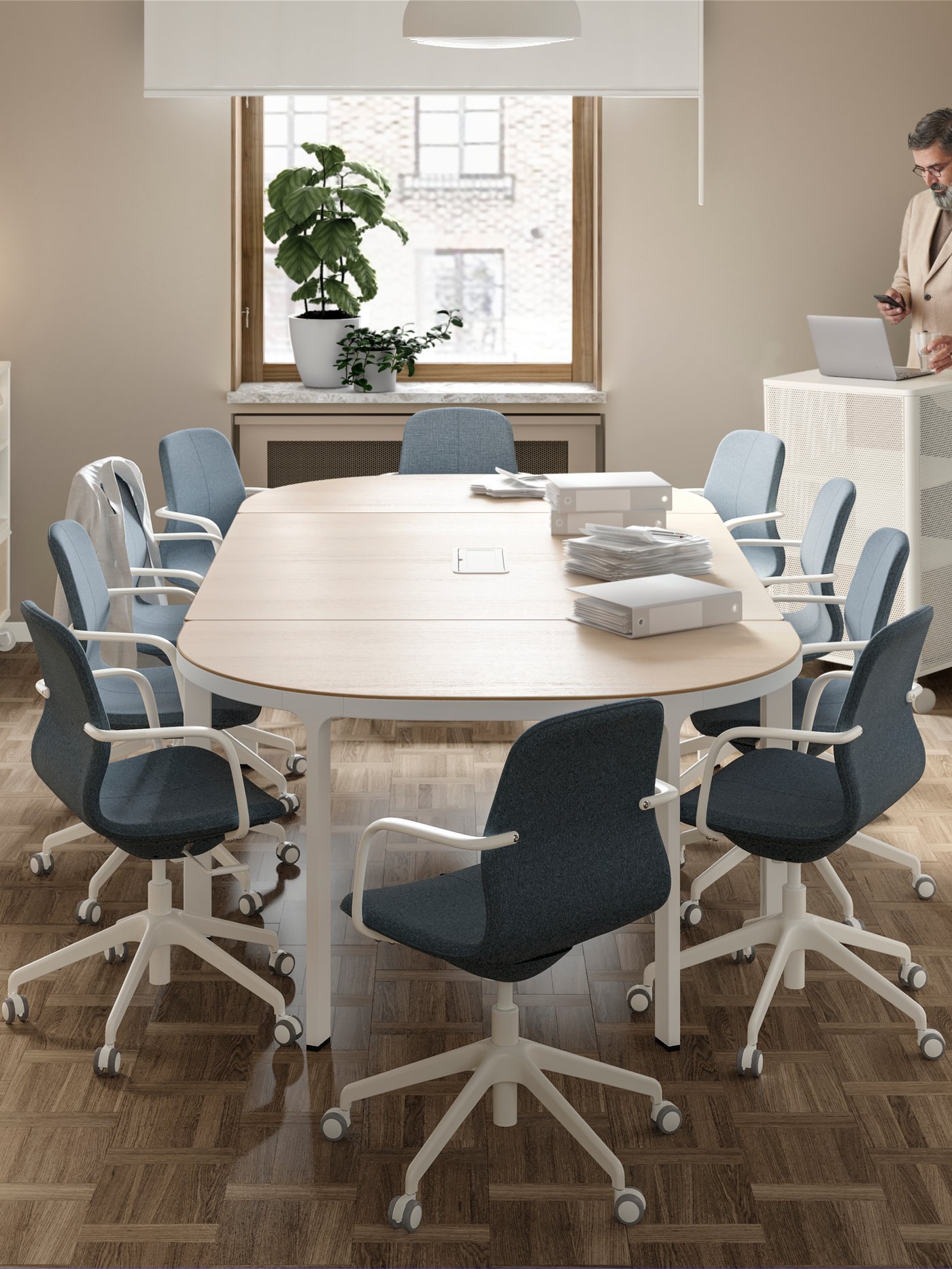 A meeting room with a conference table and chairs. At the back is a window with two potted plants and a white screen.