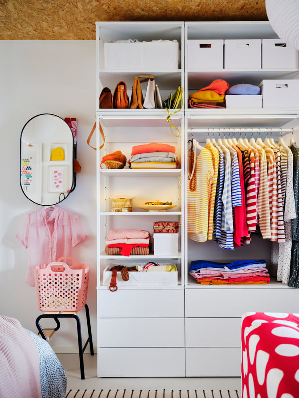 A high, white PLATSA wardrobe with a combination of closed and open storage, filled with colourful garments.