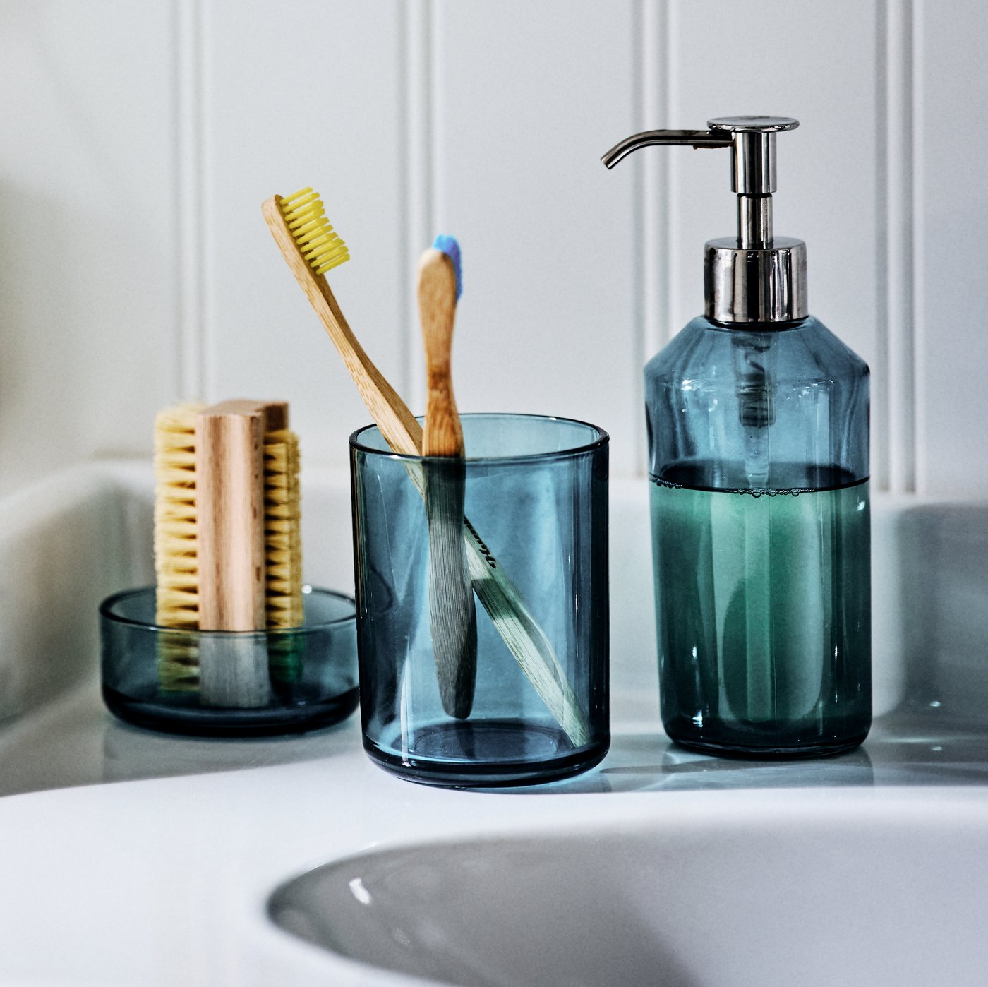 The SKISSEN glass three-piece bathroom set on the side of a wash-basin, holding a nail brush, toothbrushes and liquid soap.