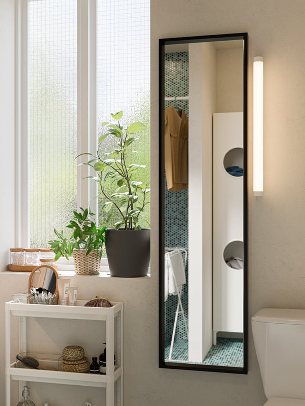 A tall and narrow black NISSEDAL mirror is mounted on a white bathroom wall. A white VESKEN trolley is standing next to it.