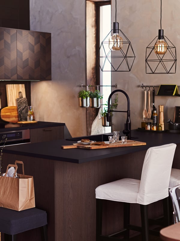 A dark wood kitchen with an island that has BRUNSTA pendant lamps overhead and glasses on the counter.