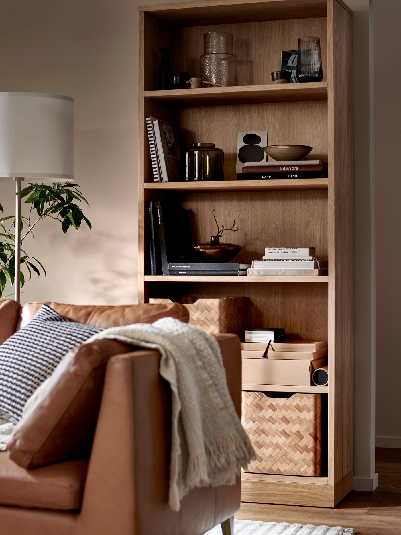 A STOCKHOLM three-seat sofa stands in front of a TONSTAD bookcase with books, vases and bamboo BULLIG boxes on its shelves.