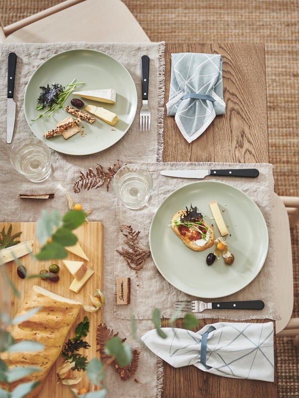 A corner of a decorated table with napkins, black LIVNÄRA cutlery, and festive food served on matt green FÄRGKLAR plates.