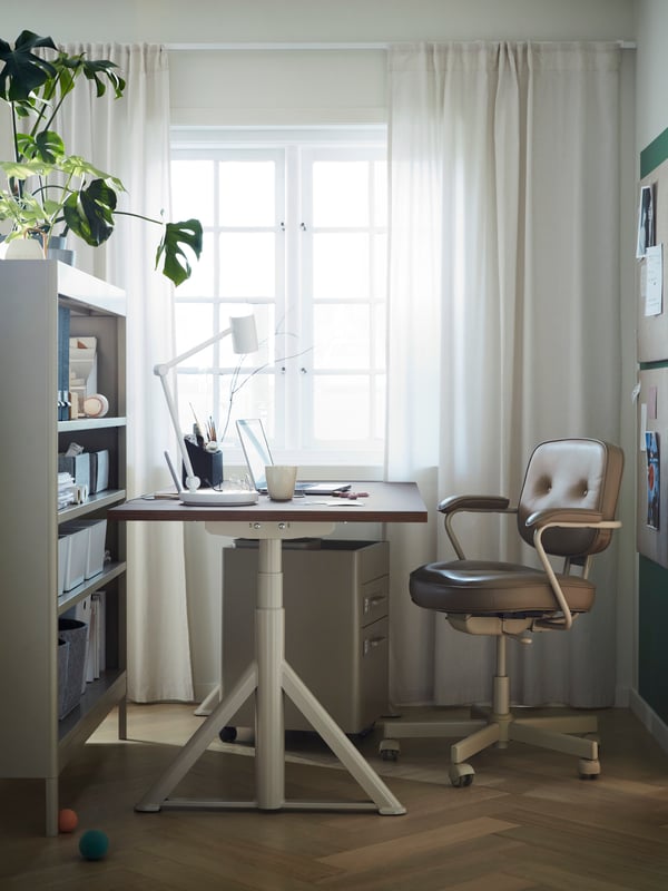 En una oficina en casa de se un escritorio de altura ajustable y una silla con apoyabrazos ALEFJÄLL.