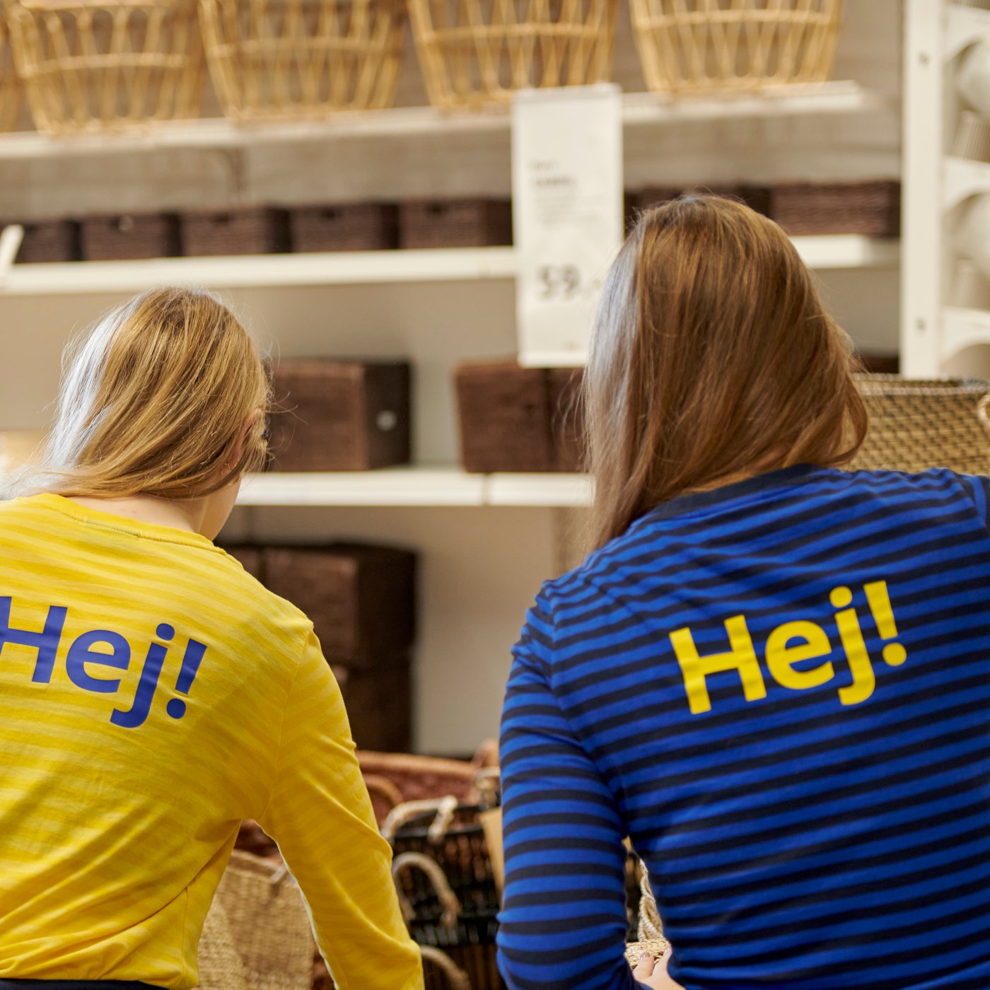 To personer står i er IKEA varhus med ryggen til. De er iført knaldblå og gule trøjer med "Hej!" trykt på bagsiden.