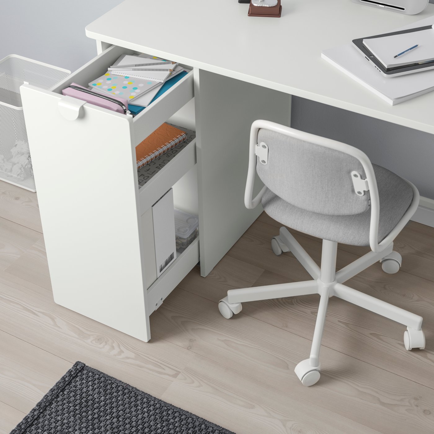 Un escritorio blanco ubicado en un rincón con muros verdes, junto al vestíbulo de una casa o departamento con piso de parquet. Frente al escritorio hay una silla de altura ajustable con asiento verde, y sobre el escritorio una repisa con artículos de trabajo.