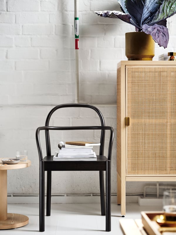 A dark brown STOCKHOLM 2025 chair with books and bound papers on it, by a white wall, between a side table and a cabinet.
