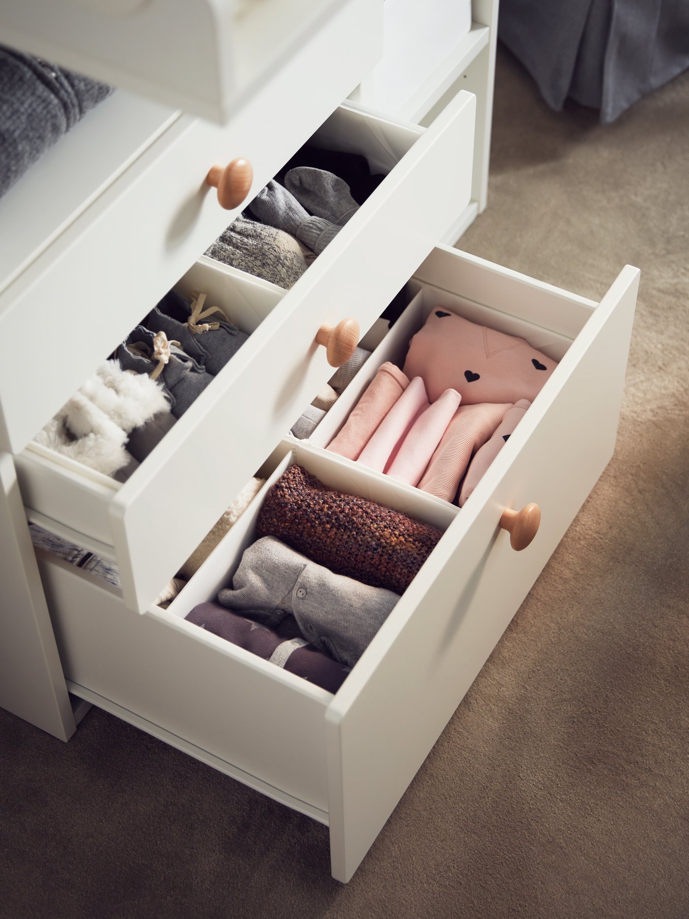 Open drawers of a changing table containing white RASSLA boxes with compartments containing neatly folded baby clothes.