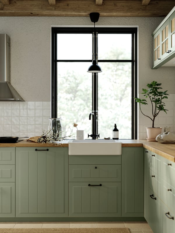 A kitchen with STENSUND kitchen fronts in light green with a window, kitchen appliances and a SKURUP pendant lamp in black.
