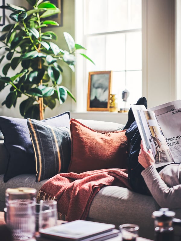 Cushions dressed in a STORTIMJAN cushion cover and DYTÅG cushion covers placed on a sofa next to a person reading a magazine.