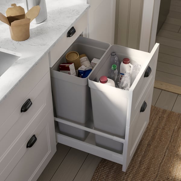 Une cuisine présentant un système blanc avec des comptoirs gris, deux bacs gris remplis de divers contenants recyclables.